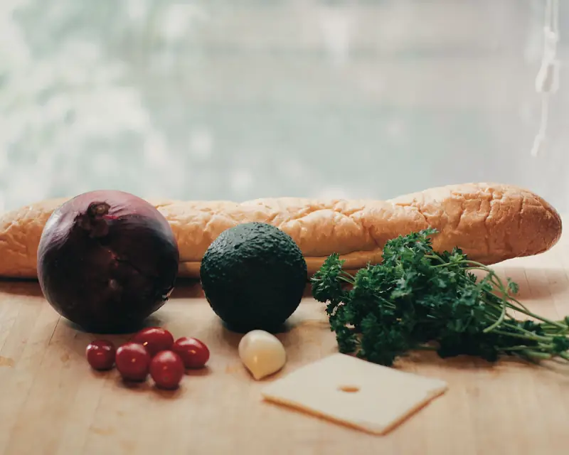 Une baguette, un oignon rouge, un avocat, des tomates cerises, du persil et une tranche de fromage sur une table en bois.